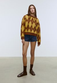 Brun och gul argyletröja med lös passform, kombinerad med jeansshorts. Modellen bär bruna strumpor och läderskor, står på ett neutralt golv.