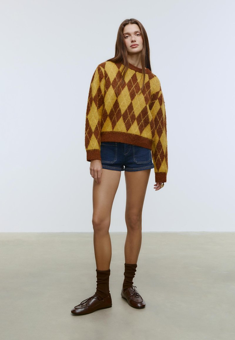 Brun och gul argyletröja med lös passform, kombinerad med jeansshorts. Modellen bär bruna strumpor och läderskor, står på ett neutralt golv.