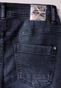 Dark denim jeans with a rear pocket, featuring a glossy label stating "embrace cosiness." The fabric has a smooth texture and minimal fading.