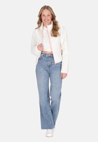 Giacca bianca trapuntata con dettagli rosa sulla zip, abbinata a jeans blu a vita alta. Il modello si trova in una posa rilassata con sneakers bianche.