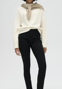 Personne portant un pull crème avec une écharpe beige nouée autour du cou et un pantalon noir ajusté, debout devant un fond clair uni.