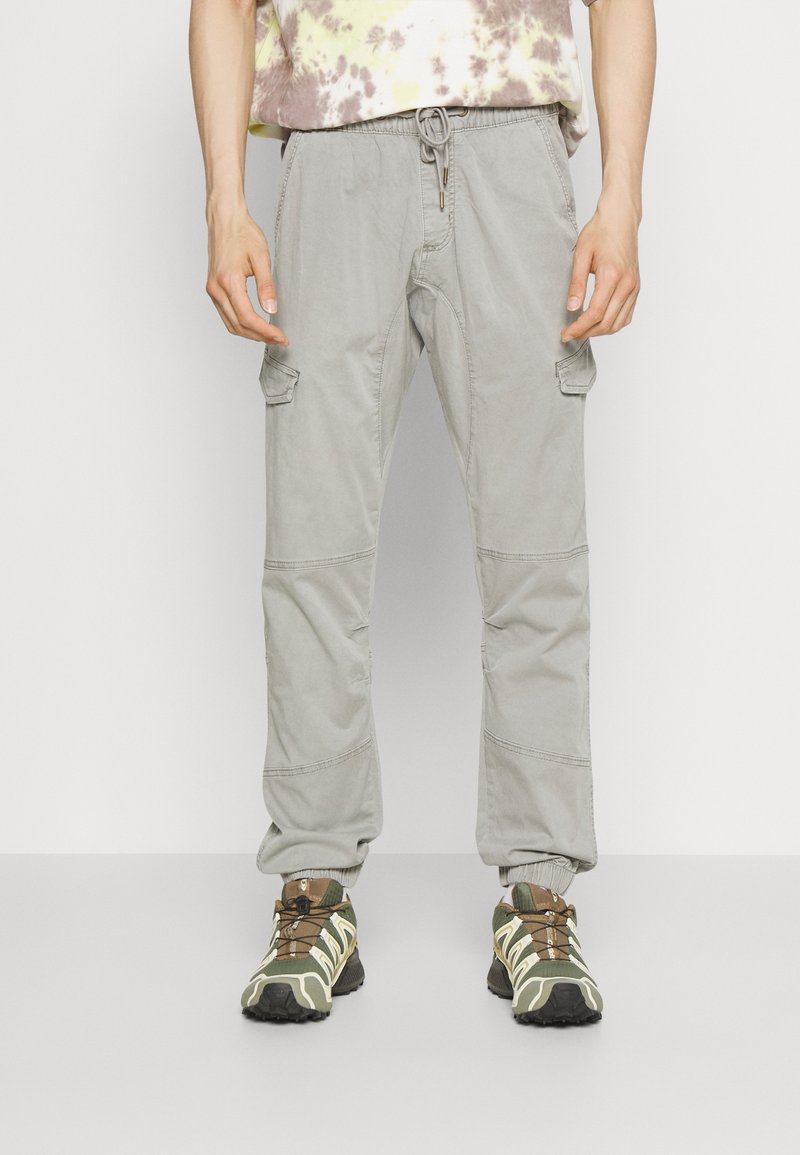 Pantalon cargo gris clair avec taille à cordon ajustable, poches latérales et coutures renforcées aux genoux, associé à des chaussures de trail vertes et marron.
