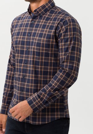 Man draagt een marineblauw en bruin geruit overhemd met knopen en lange mouwen, met borstzak, getoond van nek tot heupen tegen een effen achtergrond.