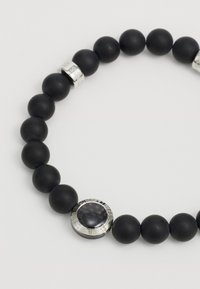Bracelet en perles noires mettant en avant des perles mates et des accents en argent. Les détails notables incluent un charm circulaire avec une surface texturée.