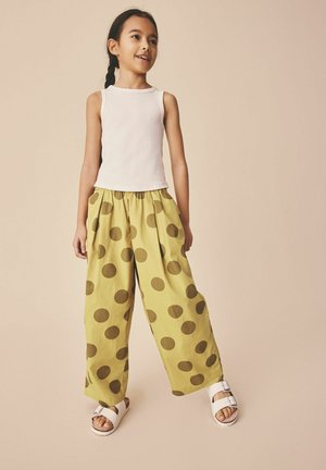 Tüdruk seisab ja naeratab, kandes valget varrukateta toppi, kollaseid laiade säärtega pükse suurte pruunide täppidega ning valgeid sandaale.