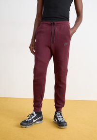 Bordo Nike trenirke s pojasom na vezanje, suženim nogavicama i kontrastnim šavovima. Imaju logo na bokovima i elegantnu, meku teksturu tkanine.