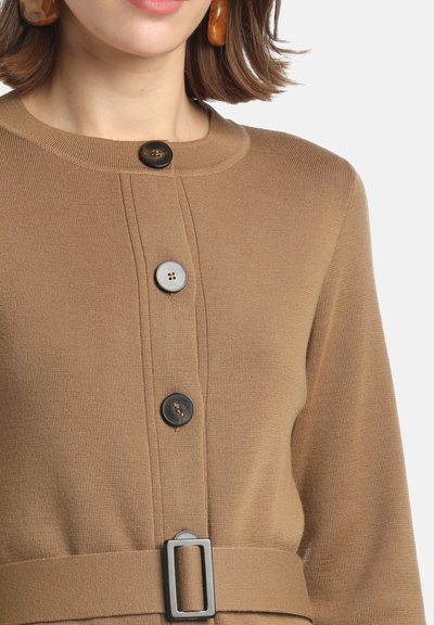 Femme portant une robe tricotée couleur camel avec des boutons noirs sur le devant, une ceinture avec une boucle rectangulaire et des boucles d'oreilles en ambre.