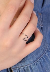 Zilveren golvende ring, dunne band, gladde afwerking. Draagt op een hand met lichte huid; denimstof op de achtergrond. Subtiele designdetails zichtbaar.