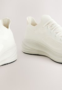 Baskets blanches de sport avec dessus en maille texturée, semelle épaisse et coussinée, et semelle extérieure en caoutchouc noir. Design minimaliste avec des accents subtils.