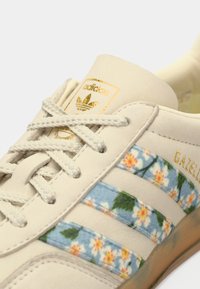 Sapatilhas de camurça creme com padrão floral em riscas azuis, apresentando atacadores beges e detalhes dourados na língua e na lateral.