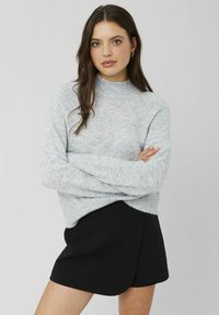 Pull en tricot surdimensionné gris clair avec un col haut et des poignets côtelés, porté avec une jupe noire de style portefeuille. Texture lisse ; coupe décontractée.