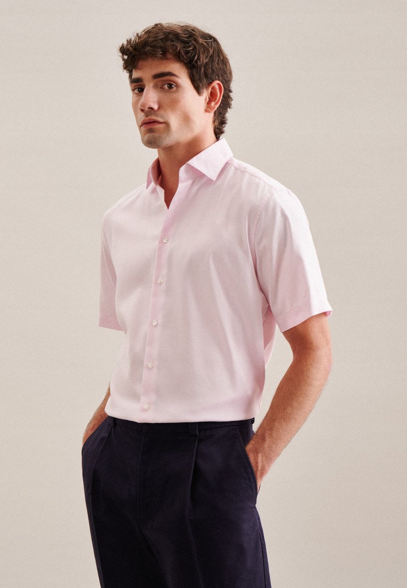 Camicia rosa chiaro a maniche corte con colletto a punta, caratterizzata da una texture liscia e bottoni perlacei, abbinata a pantaloni blu navy.