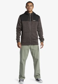 Zip-up hoodie featuring a dark gray fleece upper and brown textured lower, with front pockets and drawstring hood. Paired with light green pants.