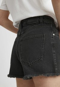 Shorts en denim noir avec un ourlet effiloché, deux poches arrière et une finition texturée. La taille est placée au-dessus de la taille naturelle.