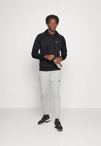 Zwarte Nike-hoodie met een voorvak, gecombineerd met lichtgrijze Nike-joggers. Model houdt een zilveren waterfles vast en draagt zwarte sportschoenen.