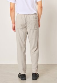 Asmuo, vilkintis smėlio spalvos kordurojų kelnes su galinėmis kišenėmis, baltą marškinėlį ir juodus sportinius batelius, stovintis ant šviesios grindų dangos priešais paprastą sieną.