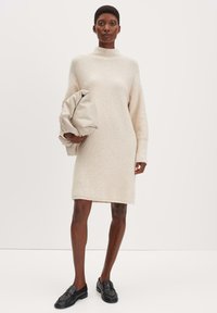 Cremfarbendes Strickkleid mit hohem Kragen, überschnittenen Schultern und langen Ärmeln. Das Model hält eine beige Clutch. Schwarze Loafers runden den Look ab.