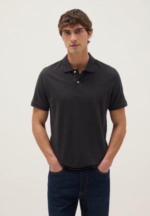 Joven con cabello castaño que lleva una camisa tipo polo negra de manga corta y vaqueros azul oscuro, de pie con las manos en los bolsillos frente a un fondo liso.