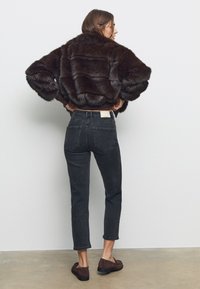 Ruskea keinoturkistakki, jossa on leveät hihat, yhdistetty korkeavyötäröisiin tummiin farkkuihin ja ruskeisiin mokkanahkaisiin liukuviihtokenkiin, nähtynä takaa.