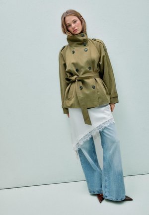 Kobieta w oliwkowo-zielonej, dwurzędowej kurtce z paskiem, białej asymetrycznej spódnicy z koronkowym obszyciem, szerokich jeansach i brązowych, spiczastych butach.