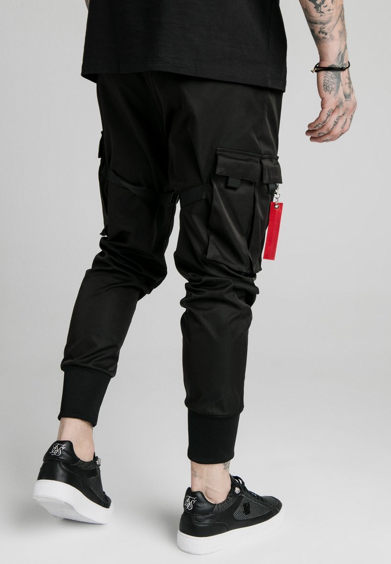 Personne portant un pantalon de jogging noir avec des poches cargo et un porte-clés rouge, des baskets noires avec des semelles blanches, et un avant-bras tatoué visible.