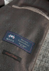 Dark brown fabric with a geometric pattern, featuring a blue label reading "Merino Lambswool" and a black label for "British Fabric."