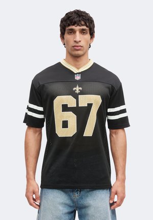Mand iført en sort New Orleans Saints NFL-trøje med nummer 67 med guld- og hvid detaljer, kombineret med lyseblå jeans.
