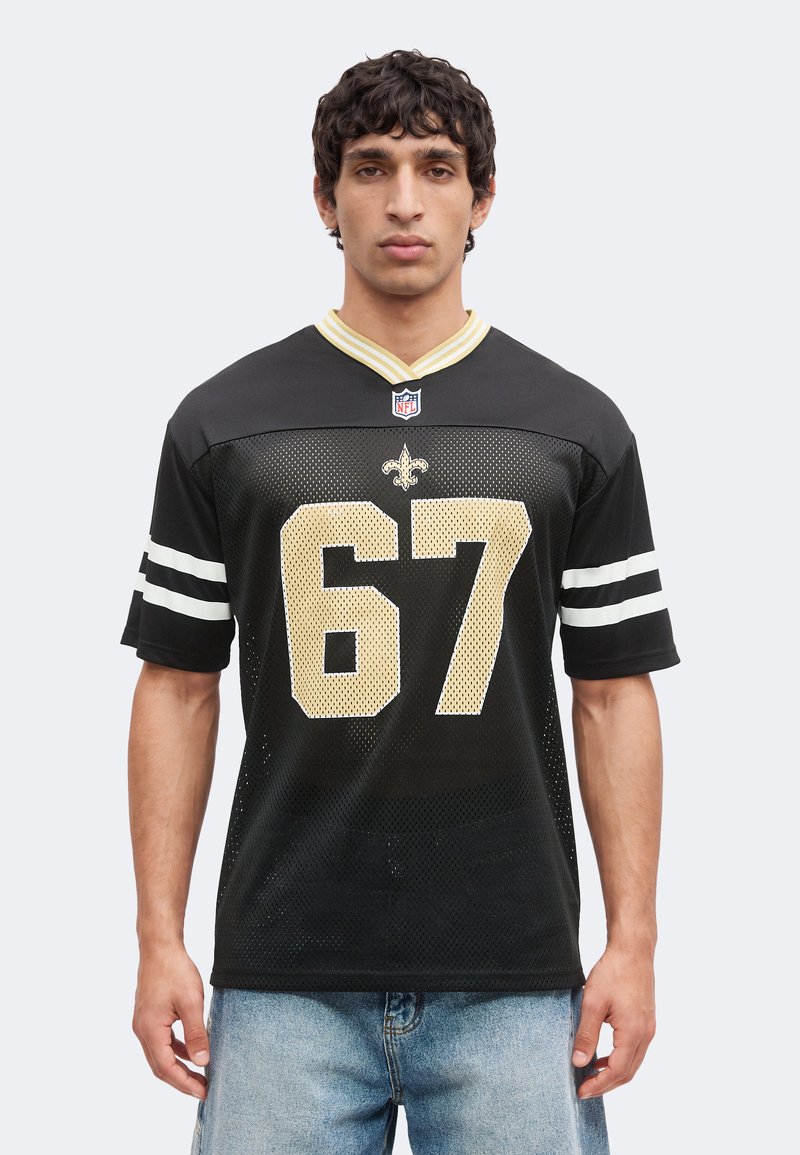 Άνδρας που φοράει μαύρη φανέλα NFL των New Orleans Saints με αριθμό 67, με χρυσές και λευκές λεπτομέρειες, σε συνδυασμό με ανοιχτό μπλε τζιν.