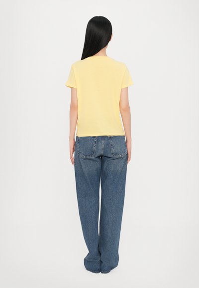 Gul kortermet t-skjorte kombinert med løstsittende, høyt liv, blå denimjeans, vist bakfra mot en hvit bakgrunn.