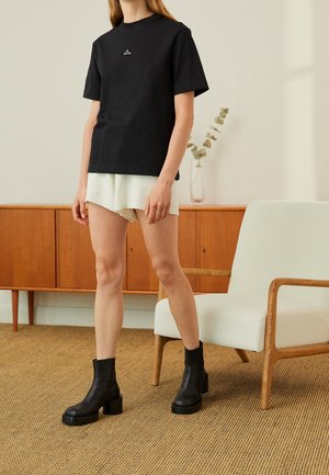 Kvinde iført en sort t-shirt med en bøjleemblem, hvide teksturerede shorts og sorte ankelstøvler med hæl står i et rum med træt gamle møbler.