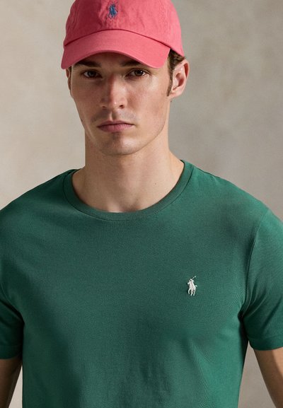 Zaļš kokvilnas T-krekls ar apaļu apkakli un mazu balta zirga logotipu. Pavadīts ar rozā cepuri, akcentējot ikdienas valkājumu.