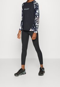 Mustat pitkähihaiset paidat, joissa on kukkakuosiset hihat, logo rinnassa; yhdistetty mustiin leggings-housuihin ja mustiin lenkkareihin, joissa on teal-väriset yksityiskohdat.