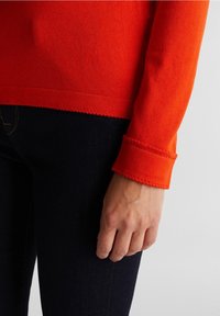 Orange knit sweater with a crew neckline and unfinished hem. The sleeve cuff features a raw edge. Worn over dark jeans, showing hand detail.