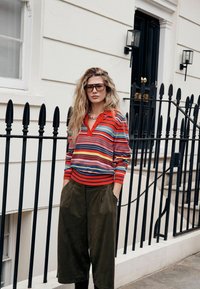 Triibuline punase ja sinise värviga pika varrukaga triiksärk kraega; oliivrohelised laiade säärtega püksid; suured prillid; tekstuuriga juuksed; linnalik keskkond musta aiaga.