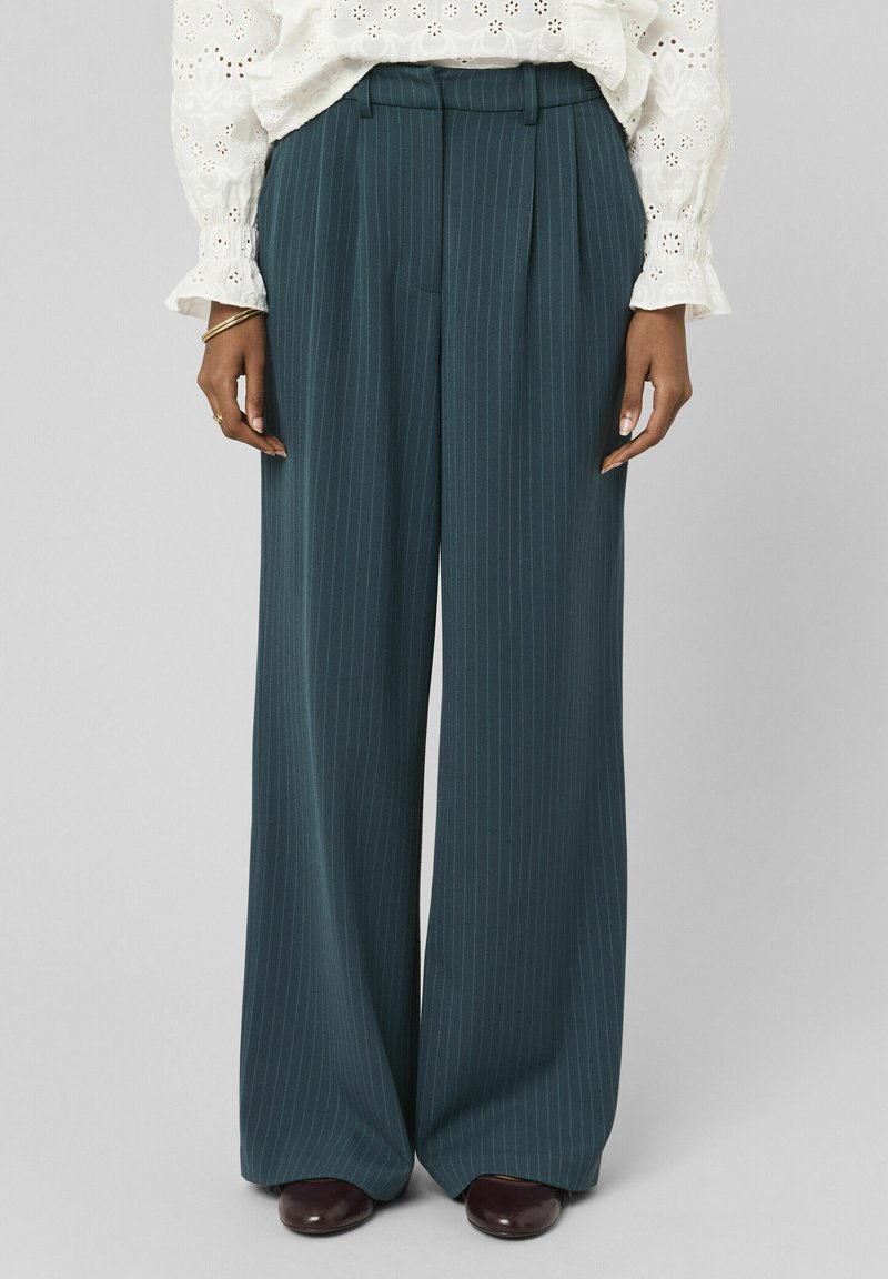 Pantalon à pinces, coupe droite, en bleu sarcelle foncé à fines rayures, porté avec un chemisier blanc en tissu ajouré et des chaussures marron foncé, visible de la taille aux pieds.