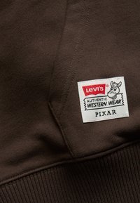 Fabric di felpa marrone con un'etichetta dai toni rossi e bianchi che recita "Levi's AUTHENTIC WESTERN WEAR PIXAR," accompagnata da una grafica di una mucca.