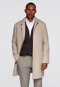 Trench coat beige sopra un gilet marrone scuro e una camicia bianca, abbinato a pantaloni grigi. Il cappotto presenta una vestibilità rilassata e un colletto classico.