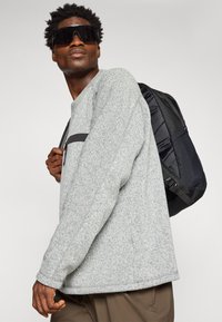 Sweatshirt texturat gri cu un buzunar de accent negru, asociat cu un rucsac negru cu un design matlasat și curele ajustabile.