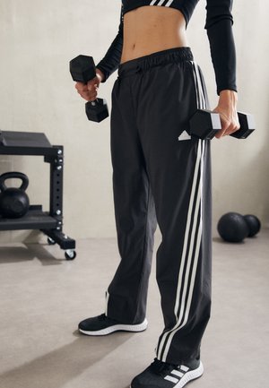 Person in schwarzen Adidas-Hosen und schwarzen Sneakers, die schwarze Kurzhanteln in einem Fitnessstudio hält, mit Kettlebell und Medizinbällen in der Nähe.