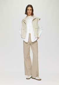 Gilet sans manches beige sur une chemise blanche, associé à un pantalon large beige clair et des baskets blanches avec des accents beige. Fond uni.
