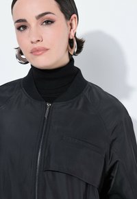 Chaqueta bomber negra con cierre frontal, cuello acanalado y un pequeño bolsillo en el pecho, con una textura suave y brillante.