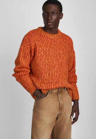 Pull en tricot orange et jaune avec une texture côtelée et un col rond, associé à un jean en denim marron clair.