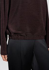 Nahaufnahme einer Person, die ein strukturiertes dunkelbraunes Langarmoberteil mit elastischem Saum und glatten schwarzen Hosen mit vorderer Naht trägt.