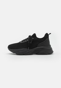 Baskets de sport noires avec une tige en tricot respirante, une semelle texturée et des lacets plats. Elles disposent d'une languette à l'arrière et d'un design sportif.