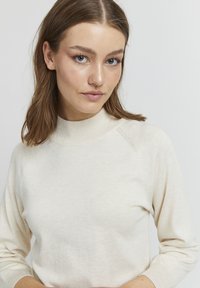 Helebeige kõrge kaelusega kootud kampsun pikkae varrukatega ja soonikute otstega. Pehme tekstuur ja peened õmbluse detailid loovad lihtsa, modernse disaini.