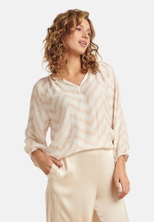 Lichtgewicht, crèmekleurige blouse met zigzagpatroon in beige. Heeft een V-hals en lange mouwen met gathered manchetten. Gecombineerd met satijnen beige broek.