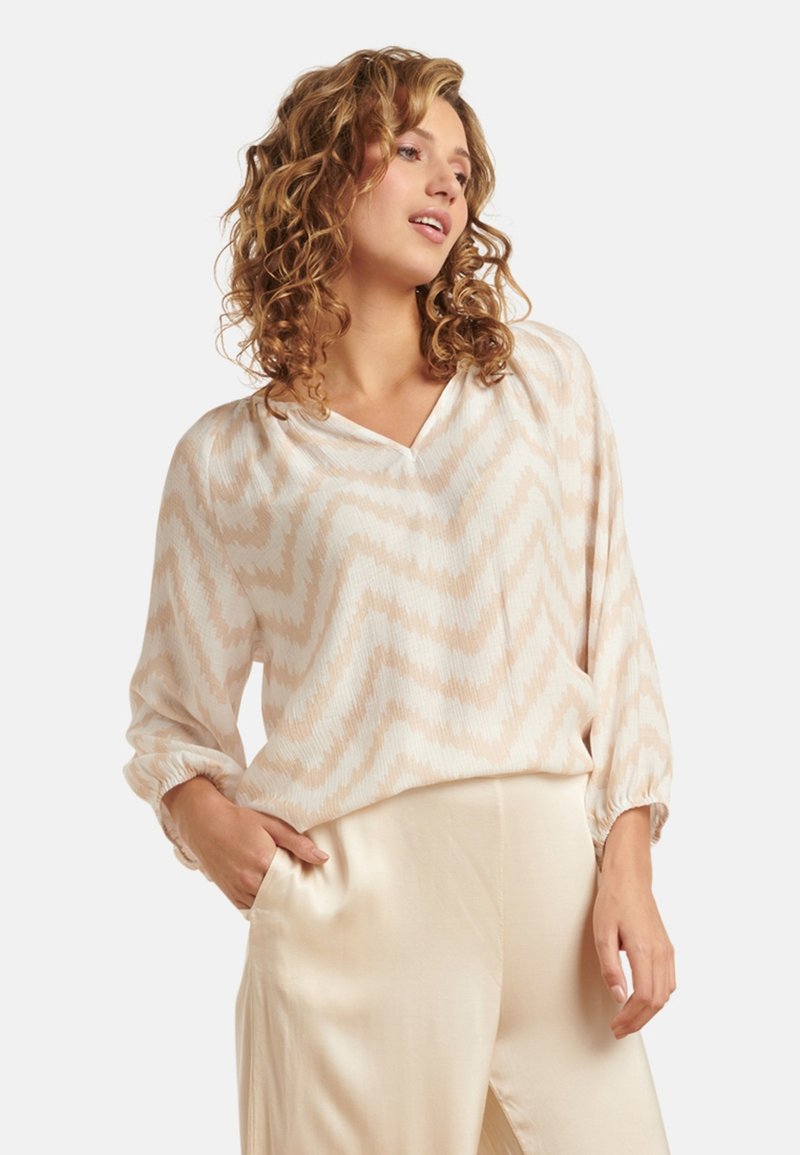 Lightweight, cream blouse with zigzag pattern in beige. Features a V-neck and long sleeves with gathered cuffs. Paired with satin beige pants.