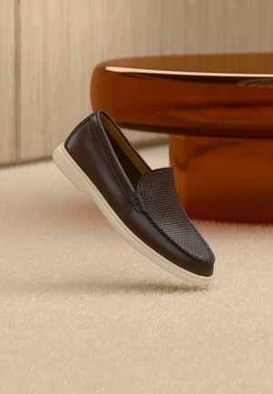 Mørkebrun læderloafer med tekstureret overdel og hvid sål, placeret på beige tæppe nær rundt, blankt brunt bord.