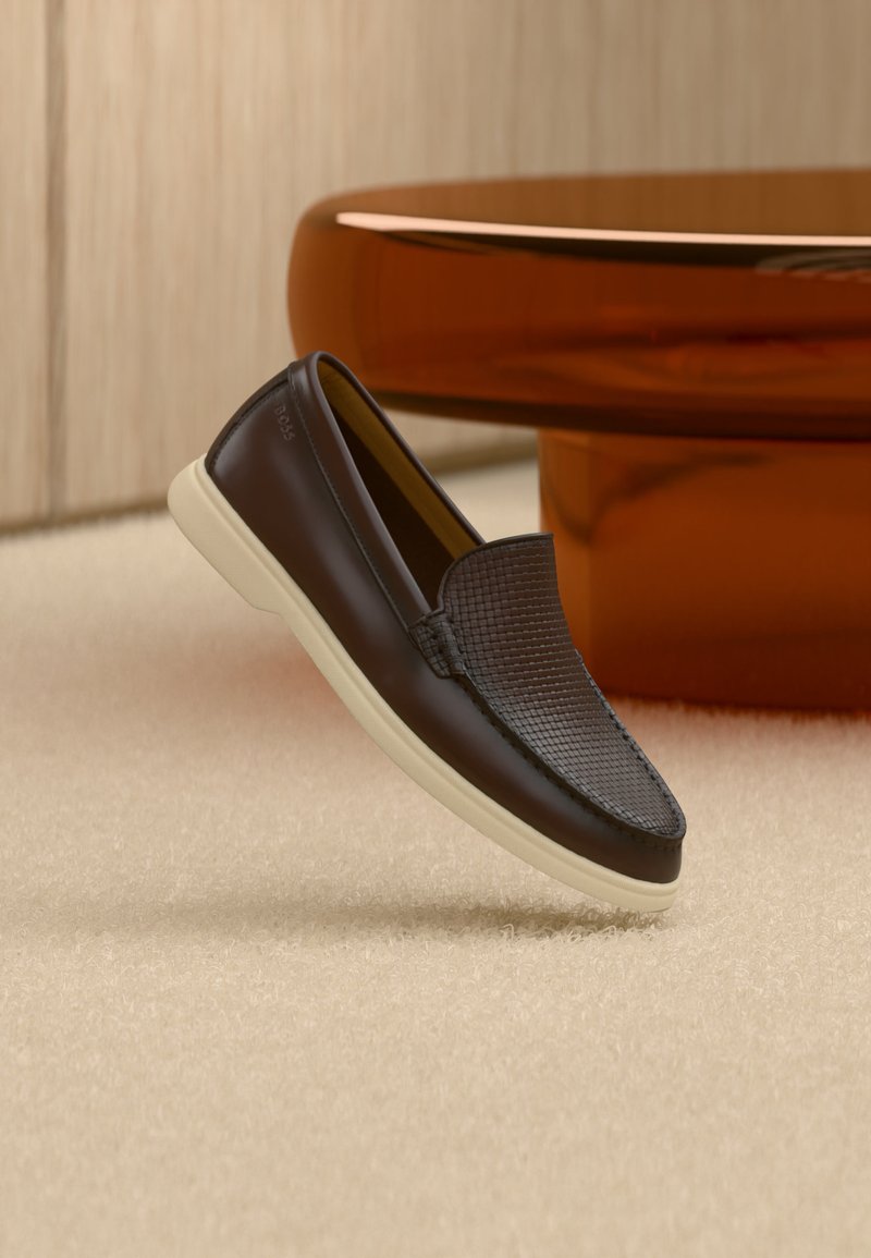 Mocassin en cuir marron foncé avec dessus texturé et semelle blanche, posé sur un tapis beige près d'une table ronde brune brillante.