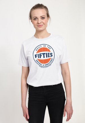 Ung kvinde med lyst hår iført en hvid T-shirt med teksten "Built in the Fifties, Original & Unrestored," iført sorte bukser, stående foran en ensfarvet baggrund.